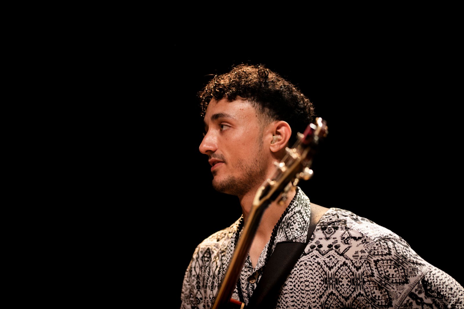 Lucas Baret in profile on stage, lit against a black ground, the neck of the hollowbody archtop crossing the frame
